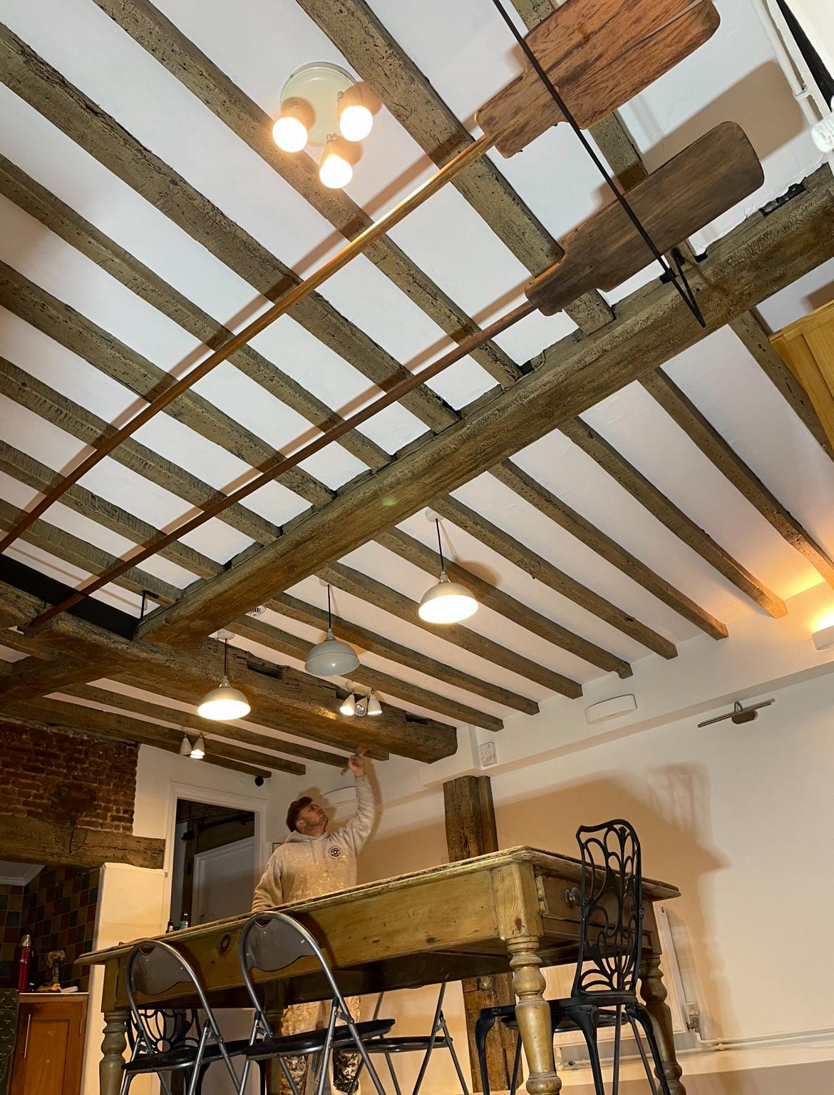 Webbs Bakery Freshly decorated beams against a white ceiling with the camera angle looking upwards. There is a large wooden farmhouse table and a decorator finishing work on the beams.