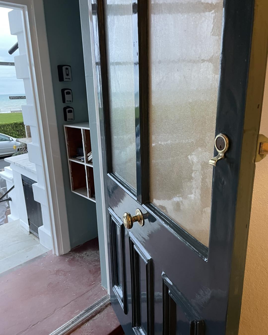 front door renovation coastal Dark glossy painted front door partially open, showing panel detailing, frosted glass panes, and brass hardware, with a coastal view visible through the doorway.
